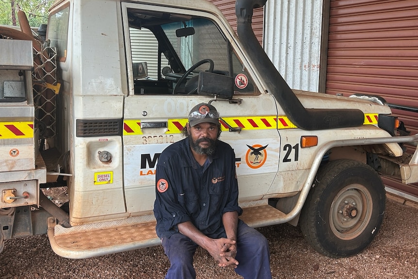 An Indigenous ranger wearing a uniform and resting alongside a work ute, which is coated in red dirt.