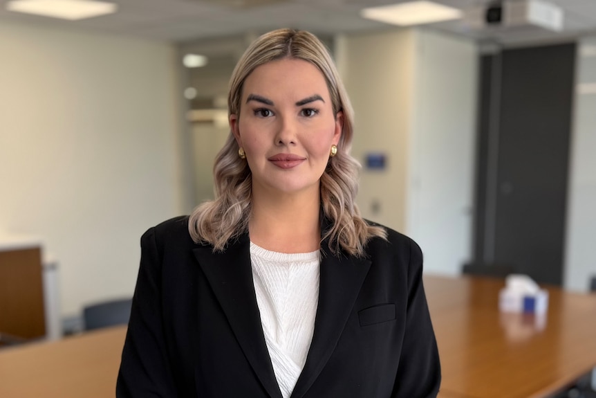 A woman with blonde hair wearing a black suit is standing infront of a boardroom table
