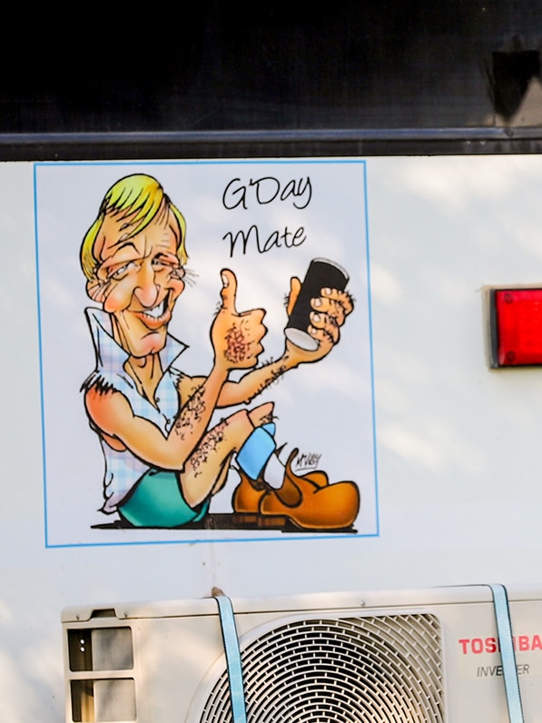 A large white vehicle sits in a caravan park it has a caricature of a man with blonde hair sitting above an airconditioner.
