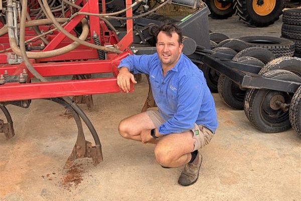 A man in a blue shirt and short squats down next to a piece of red farm machinery.