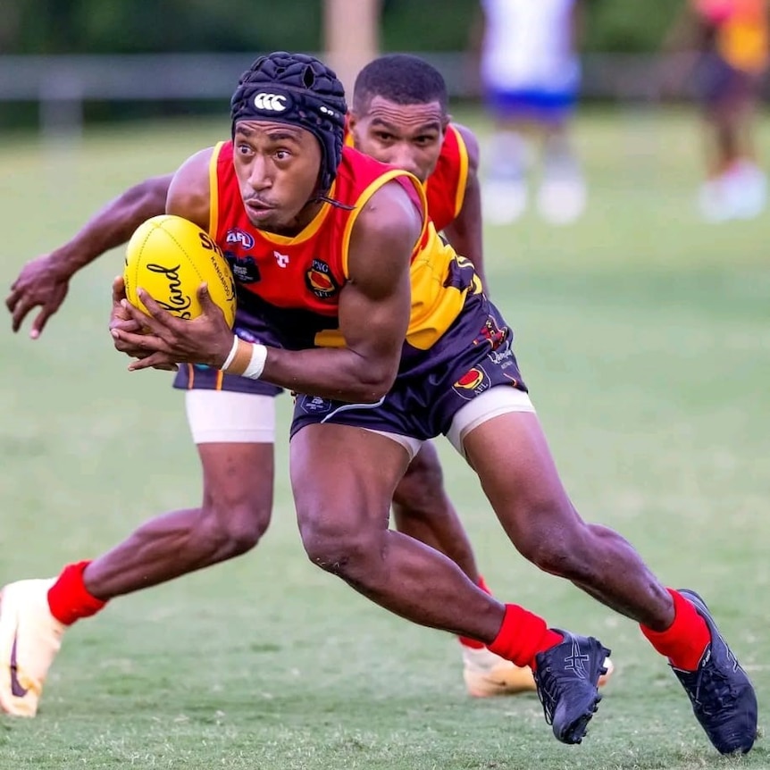 A young PNG man wearing black head gear and wearing PNG AFL uniform holds a yellow ball while playing on a field
