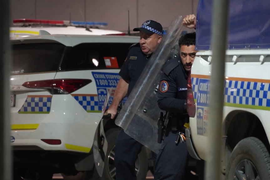 Two police officers with a riot shield behind a police vehicle.