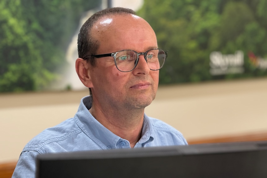 A man wearing glasses sits looking at a computer with the top of the screen just in frame.