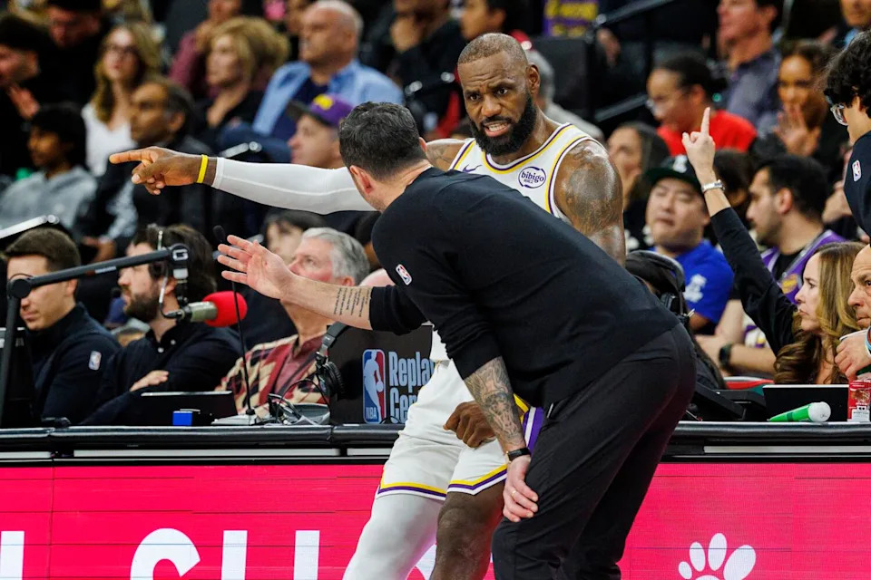 LeBron James sits on the scorer's table as he chats with Lakers coach JJ Redick during a game.