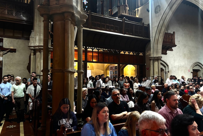 Full church pews and several rows of people stand at the back of a cathedral