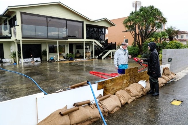 A man stands in front of his house which he has boarded up with sandbags.