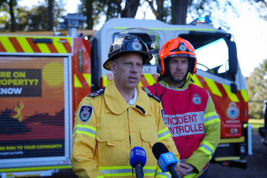 Commissioner of the Rural Fire Service Trent Curtin in uniform at a press conference, flagged by a colleague behind him.