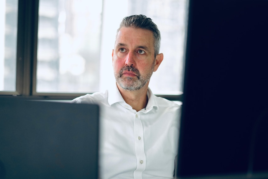 A man with a salt-and-pepper beard sits behind computer screens in an office.