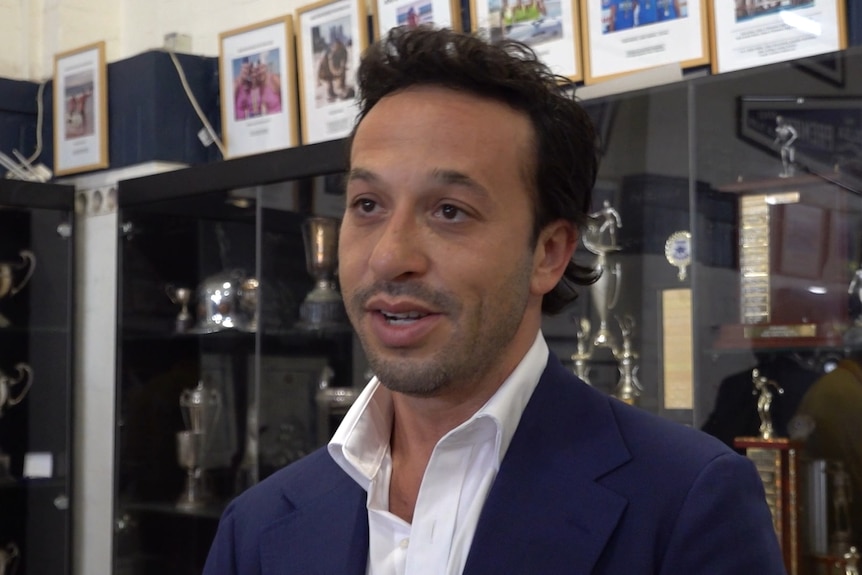 Mr Zizer in a blue blazer speaks to the camera as he stands in front of a cabinet of trophies at the Bondi surf club.