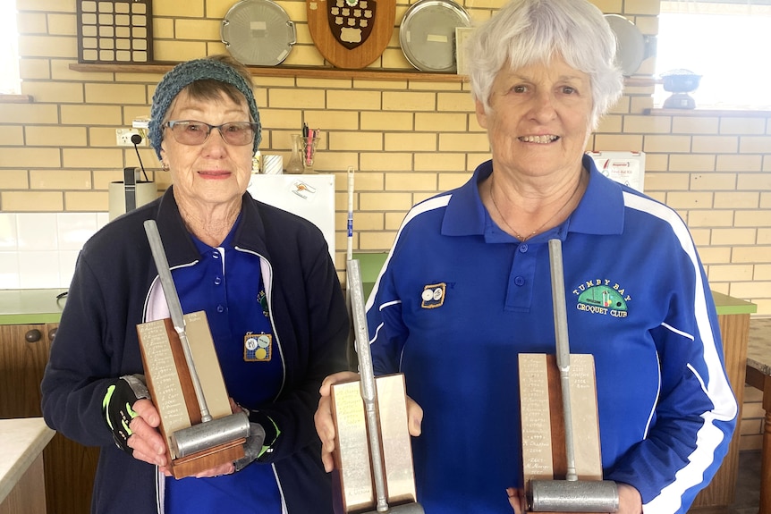 Two women holding up trophies, standing in front of wall with a shelf of shields