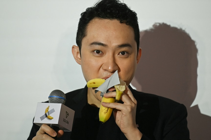 A man holding a microphone in one hand eats a banana with duck-tape hanging from it in the other