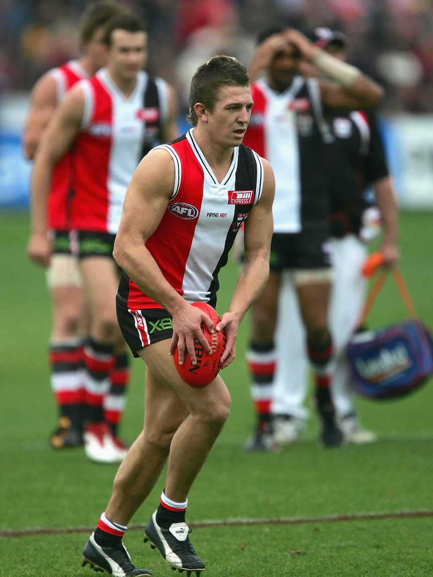 A St Kilda AFL player holds the ball as he prepares to kick for goal at the end of a game, as his teammates watch on.