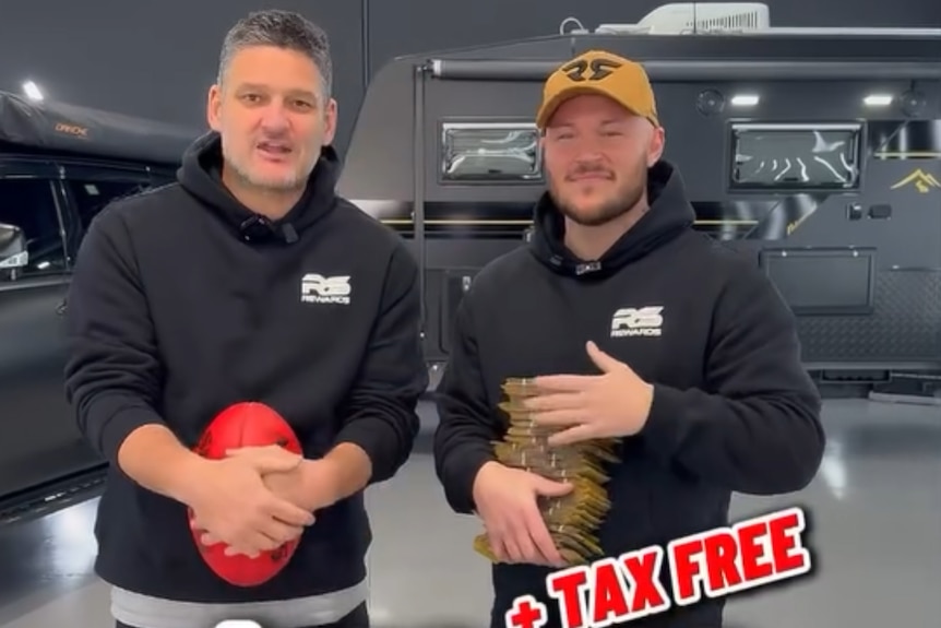 Two men in identical branded sweatshirts standing side-by-side, one holding a red football.
