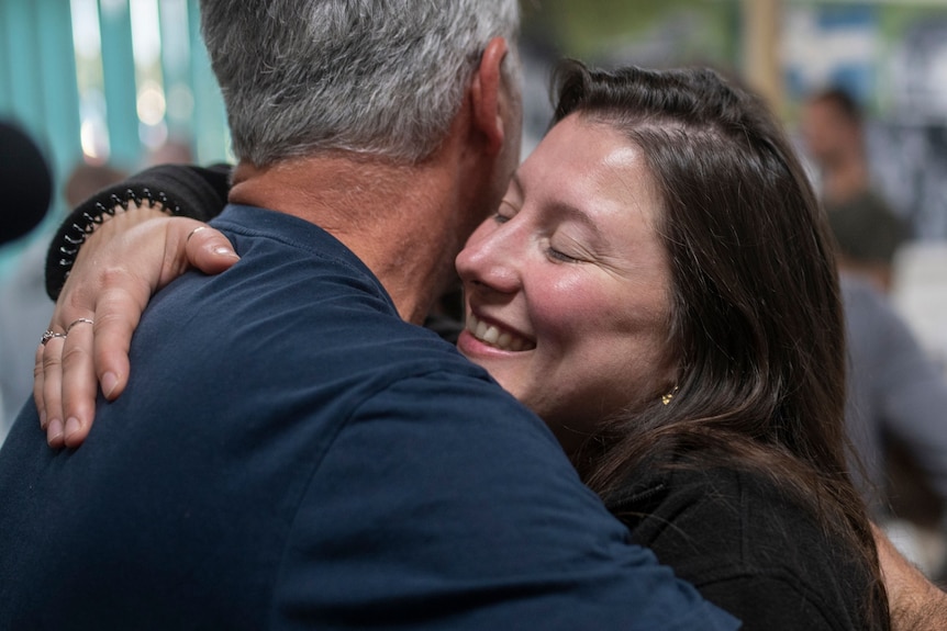 A woman with brunette hair hugs a man who has grey hair and wears a navy T-shirt.
