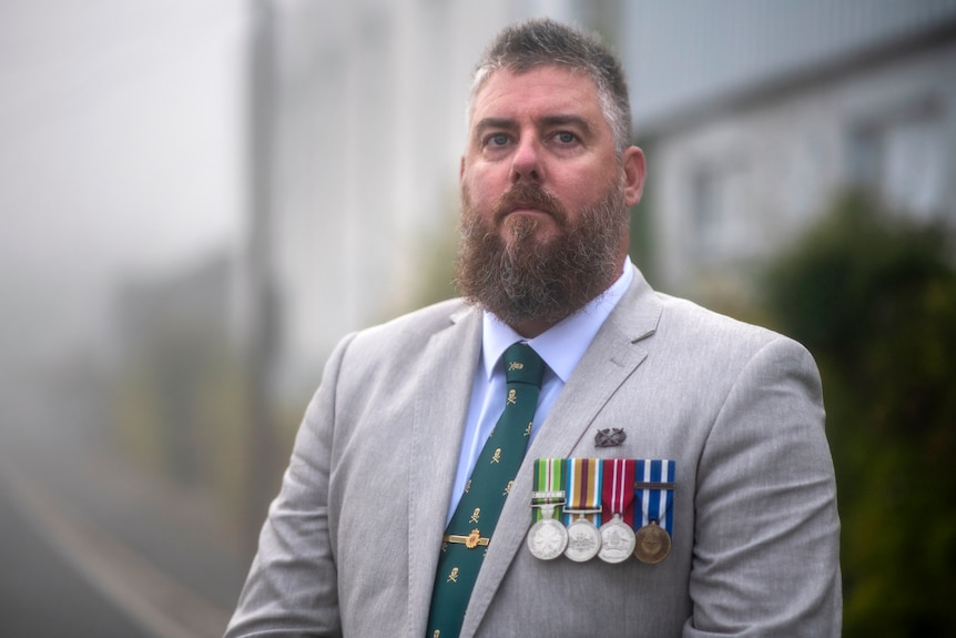 Man with beard wearing light-coloured suit with medal attached looks serious.