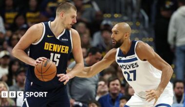 Nikola Jokic holds a basketball and stands in front of Rudy Gobert