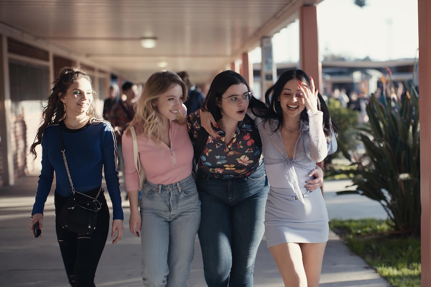 Four teen girls walk down a outdoor hallway with their arms around each other.