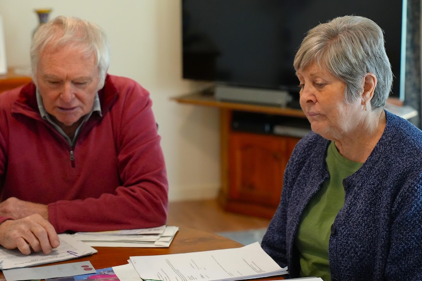 An older man in a red jumper and a older lady in a blue cardigan looking at documents on a table.