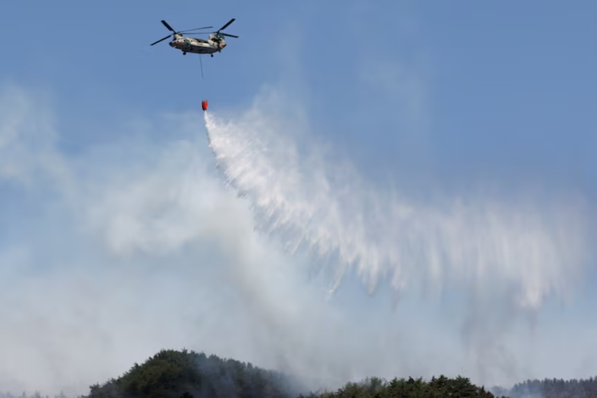 A helicopter in the blue sky drops fire suppressant over a mountain.