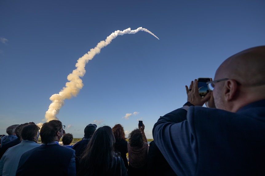 A crowd looks up in the early morning at a massive contrail from a rocket that is now pinprick in the blue sky.