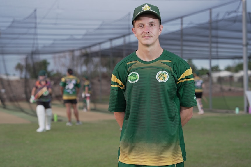 A man dressed in a green and gold cricket uniform, standing in front of players training at the batting nets.