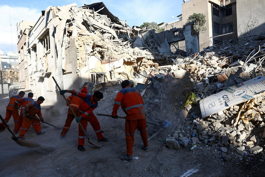 Men in orange worksuits shovel rubble by a collapsed synagogue.