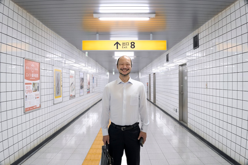 A smiling man stands in a brightly lit train station hallway