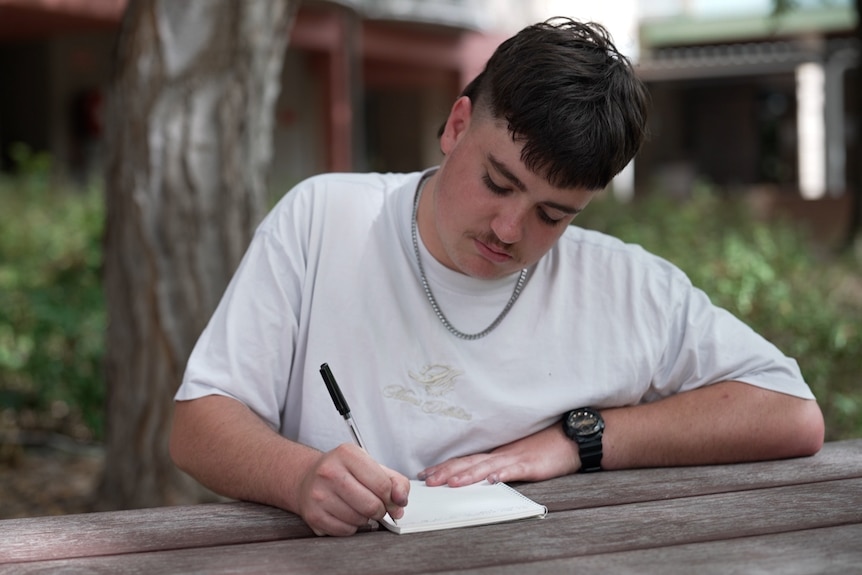 A young man writes on a bench. 