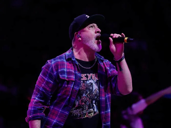 A man performing on stage, wearing a flannel shirt over a graphic tee and a cap, singing into a microphone