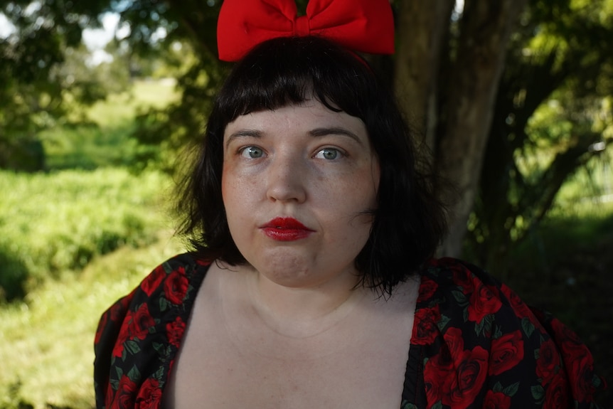 A woman with dark hair and a red bowtie in her hair stands below a tree