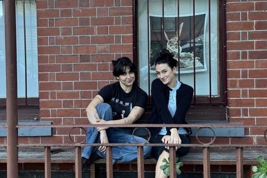 Two young women with dark hair smiling on a bench in the front of their terrace hosue.