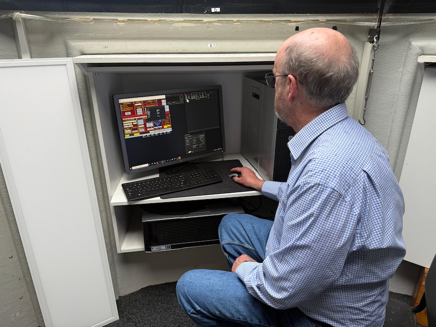 A man wearing a blue checkered shirt sitting down, using a mouse on a computer inside