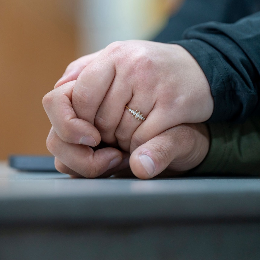 Two hands lean on a table intertwined holding each other. A female hand on top has a ring on one of the fingers.
