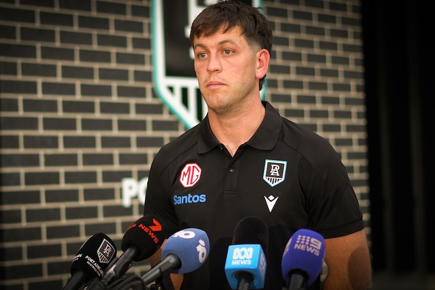 A dark-haired young man in a dark polo shirt faces the media outside a sports club.