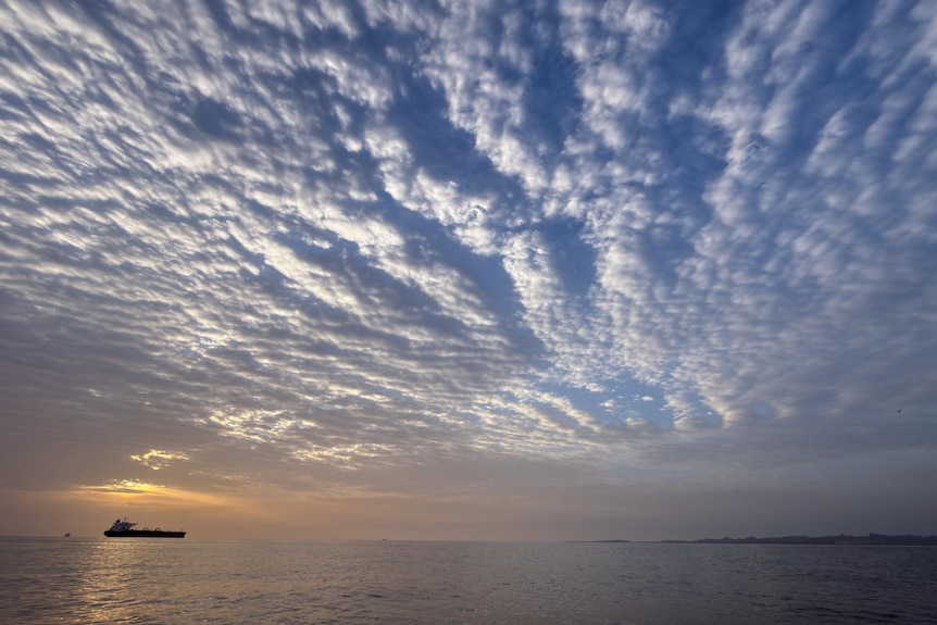 The sun rises behind a ship sitting on sea with puffy clouds above