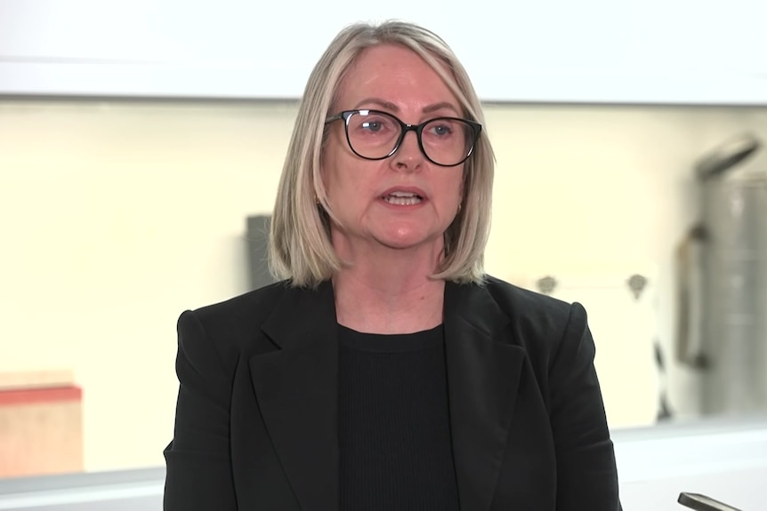 A woman with blonde hair and black glasses wearing a black jacket and top inside