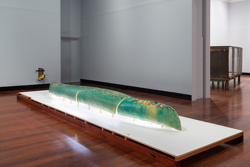 In a white gallery, an upturned canoe is mounted on a platform. The canoe has a patina like crocodile skin, using beeswax.
