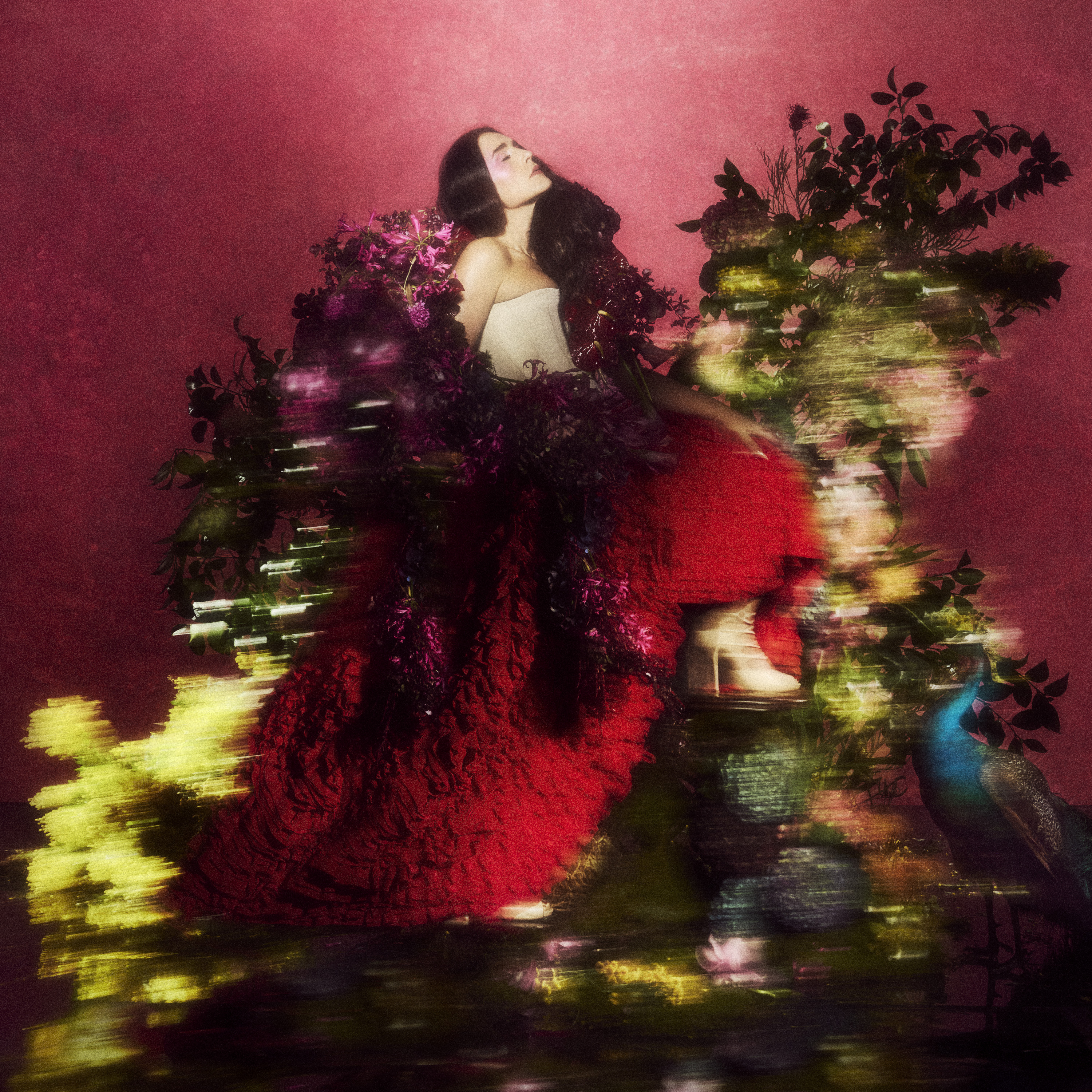 A brunette woman in elaborate white and frilly red dress poses in front of a large, colourful floral arrangement
