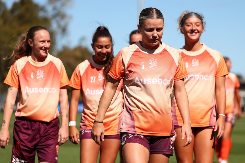 A group of women in orange shirts walk across a football field. 