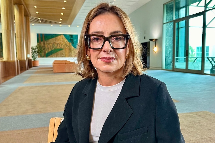 A woman with a light brown bob and thick square glasses wearing a navy blazer and white shirt sits in a large, empty foyer.