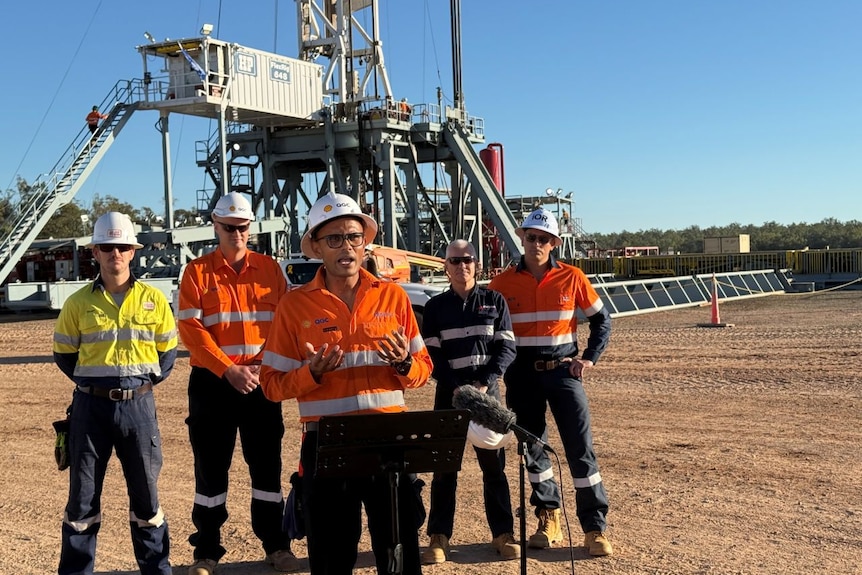 A man wearing a hard hat and a hi-vis shirt speaks to the media in a disty field.