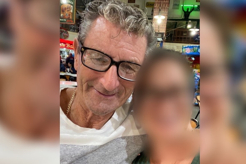 A man wearing glasses stands next to a person with a blurred face.