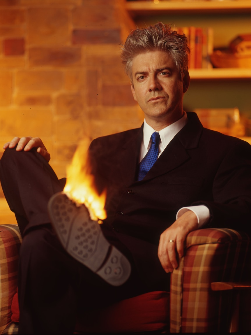 Shaun Micallef, late 30s, in a suit and tie, sits in a chair, one shoe seemingly on fire.