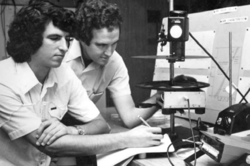 A black and white photo of two men with shoulder-length hair in a lab.