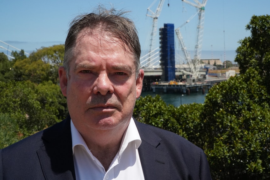 A man in a suit with a solemn expression, behind him is a port with cranes