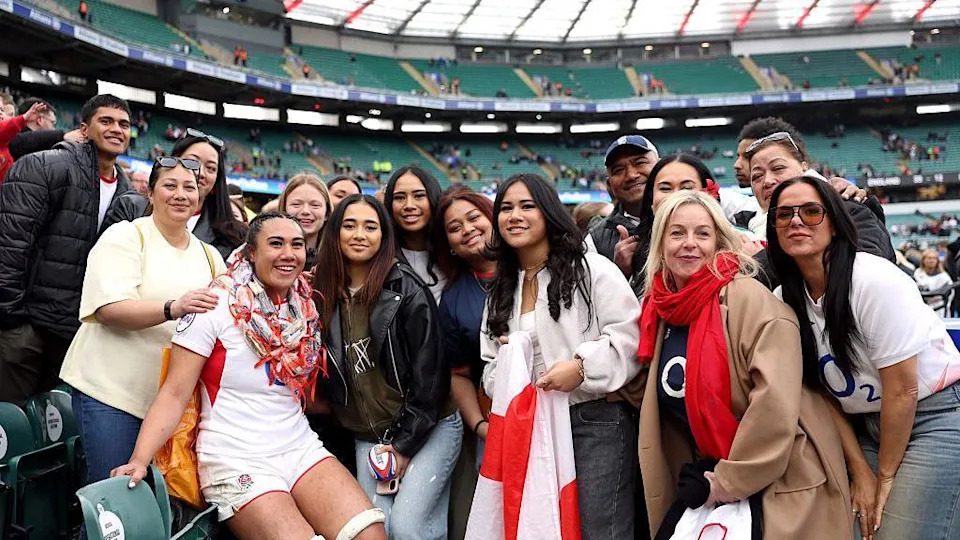Haineala Lutui with family and friends. She is wearing the England kit and a woman at the front has a flag. The group are smiling towards the camera. 
