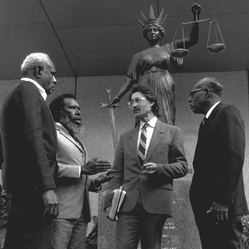 Eddie stands with a group of people, a statue of the scales of justice in the background.