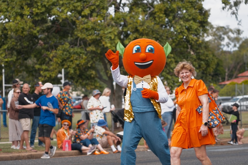 A person dressed up as an orange mascot walks along a parade next to Dael who wears an orange dress.