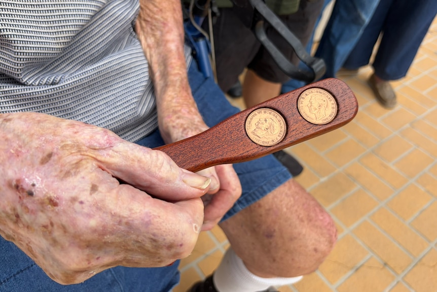 An elderly hand holds a small woodern plank with two copper coins on it