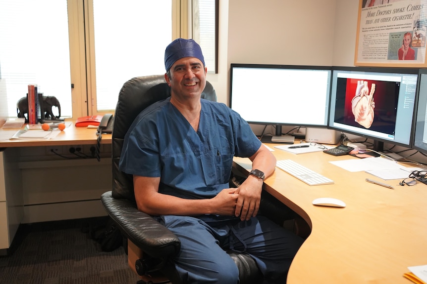 A man in navy surgical scrubs sits in front of a computer and smiles.